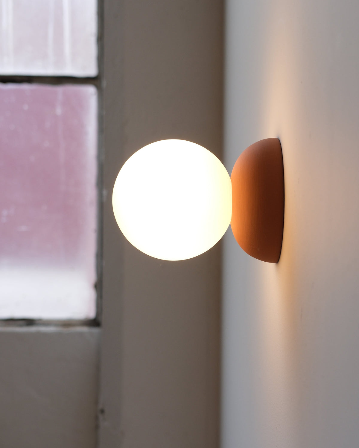 Monroe Sconce in Terracotta, lit, beside a window