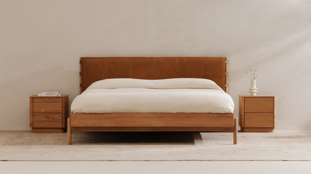 Moe's Home Collection Colby bed with a tan leather headboard and oak frame, made up in cream linen between two oak two-drawer nightstands against a pale plaster wall