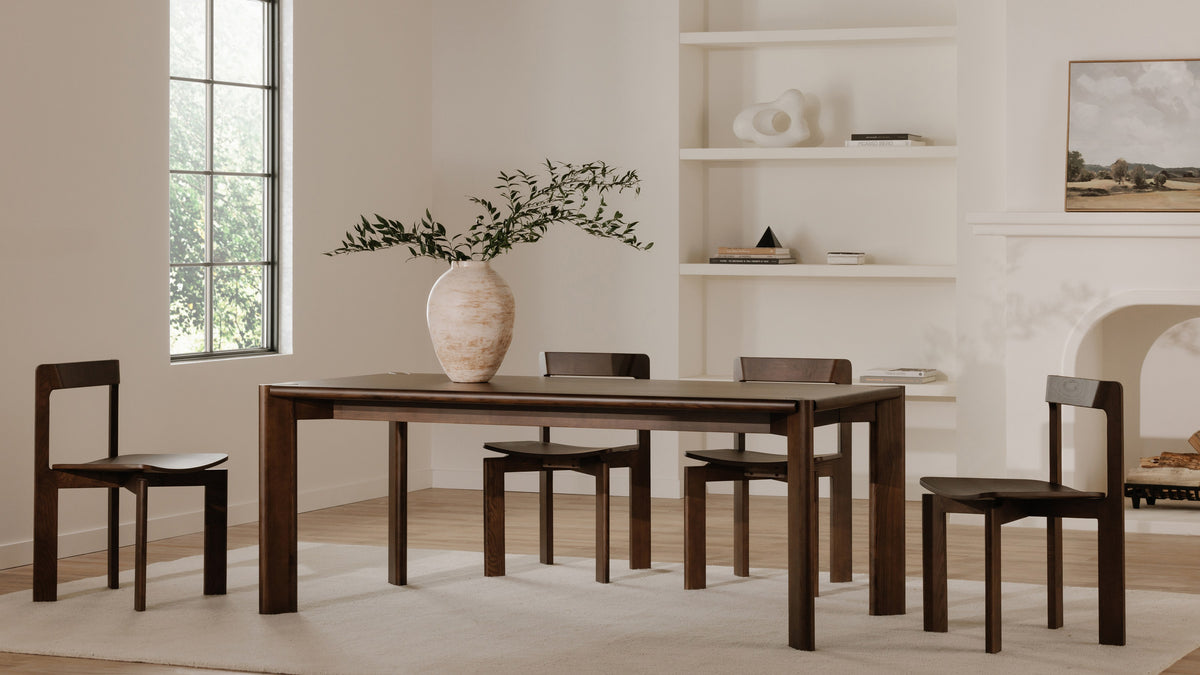 Four Moe's Home Collection Daifuku dining chairs in walnut-stained ash around the matching Daifuku dining table, in a pale room with a plaster fireplace, open shelving and a tall window