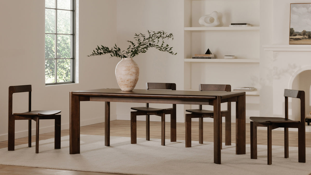 Moe's Home Collection Daifuku dining table in walnut stained ash in a bright dining room, with four dark wood chairs, a cream vase of olive branches, a tall window and open shelving behind
