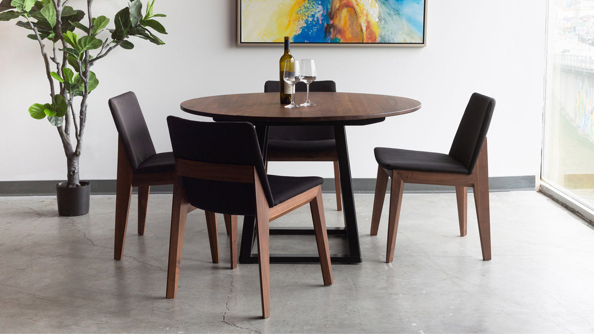 Four Moe's Home Collection Deco dining chairs in black fabric around a round walnut table on a concrete floor, with a fiddle-leaf fig and an abstract painting behind