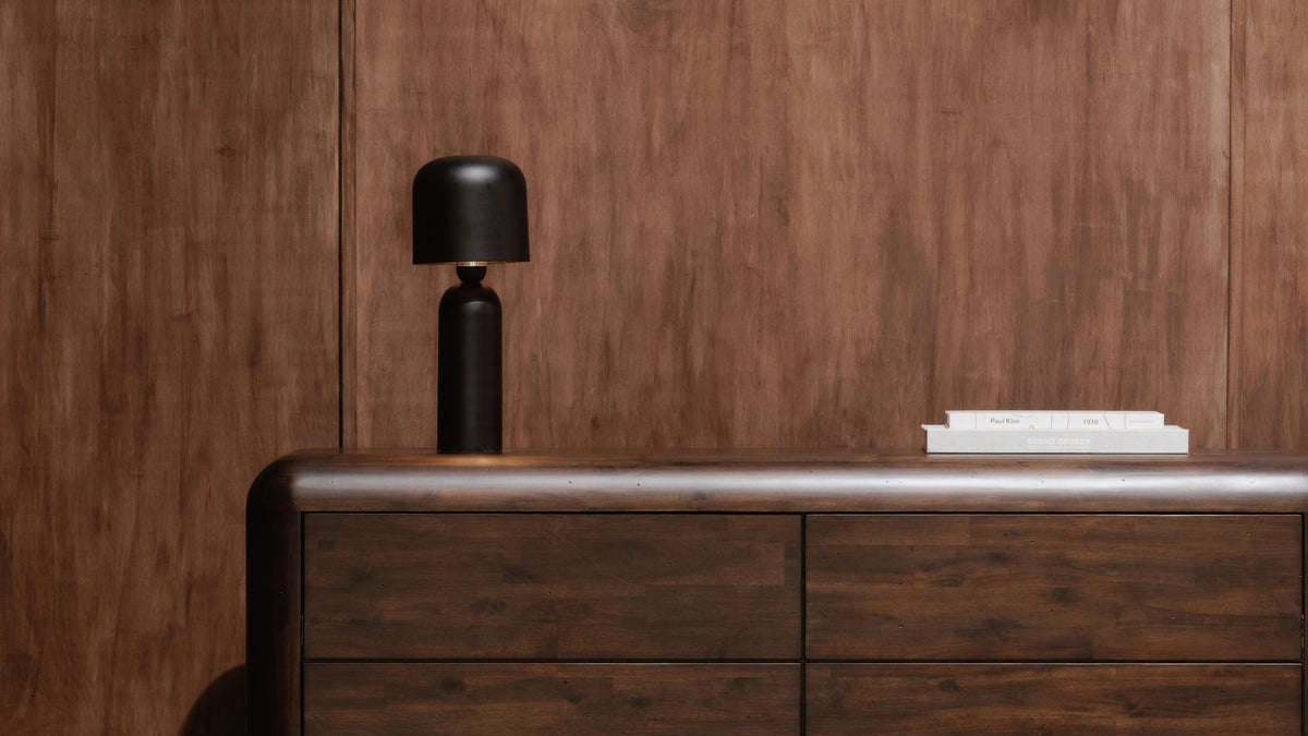 The Echo table lamp in black standing on a dark walnut dresser against a wood-panelled wall, a stack of white art books to one side
