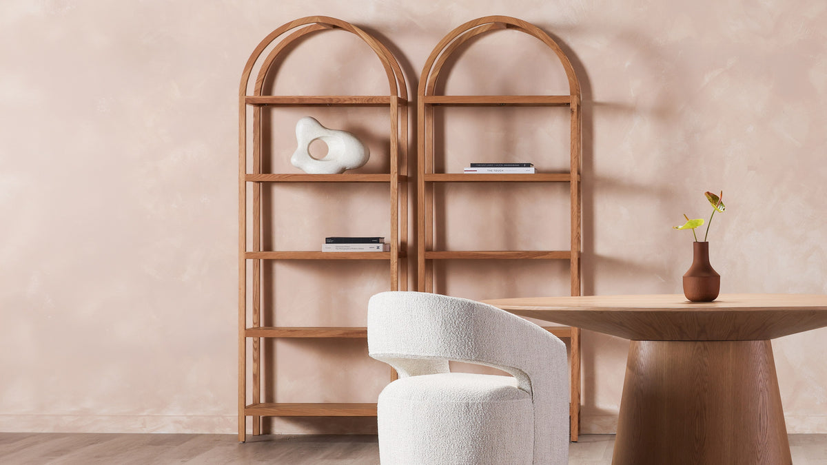 Two Moe's Home Collection Eero bookcases in natural oak standing side by side behind a round table and a boucle chair, their arched frames reading as one run