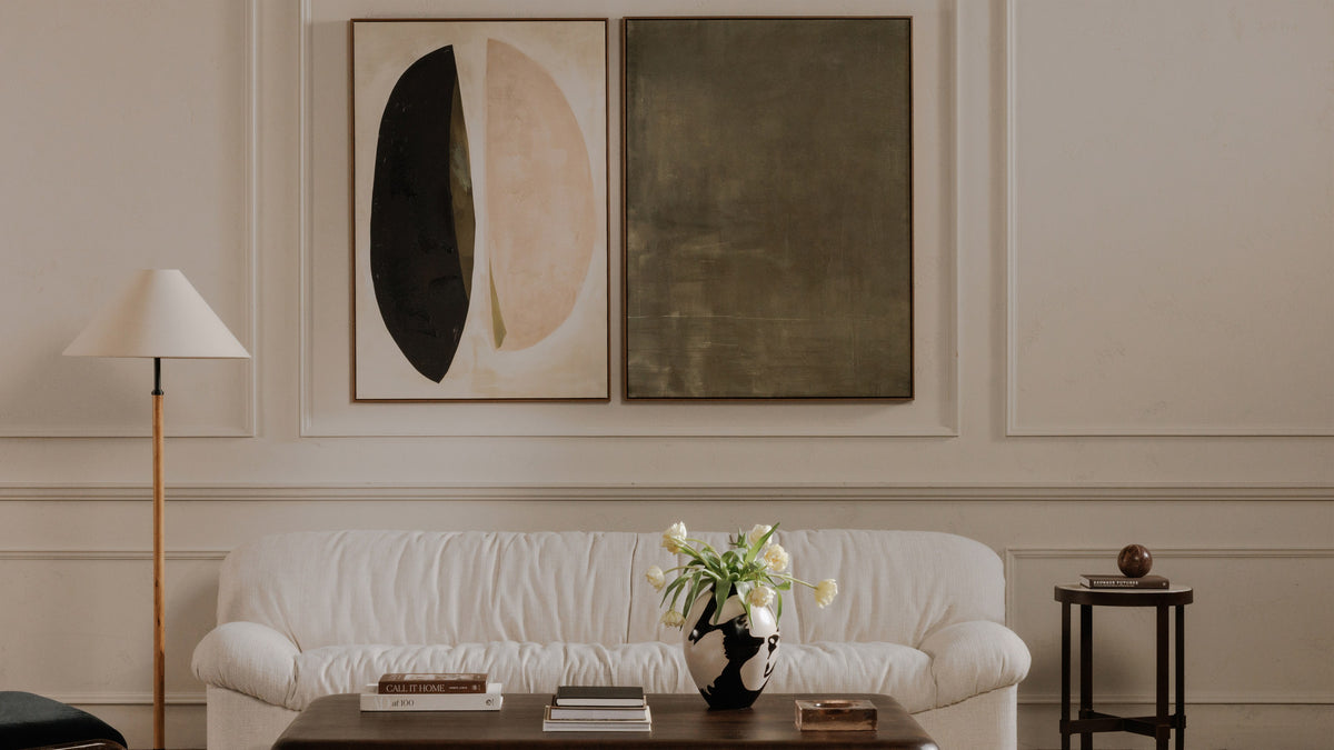 The Enamor framed painting in olive hung beside a second canvas on a paneled wall, above a white sofa with a floor lamp and a low table of books