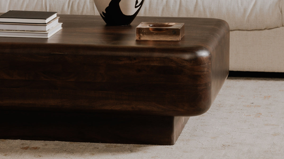 Close view of one rounded corner of the Eton coffee table, showing the thick dark acacia top overhanging its recessed plinth on both faces, with a stack of books, a black and white vase and a copper tray on the surface