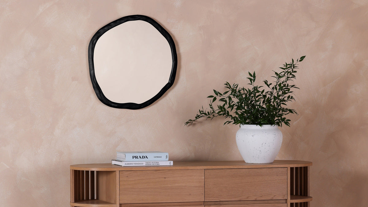 The small Foundry mirror in black on a warm pink plaster wall above a slatted wood credenza, next to a white pot of trailing green leaves