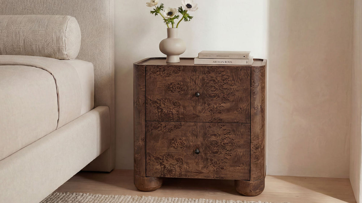 Moe's Home Collection Gibson nightstand in dark brown burl beside an upholstered bed, its two drawers and small dark knobs below a lipped top holding white anemones in a cream vase and two books, standing on rounded feet against a pale plaster wall