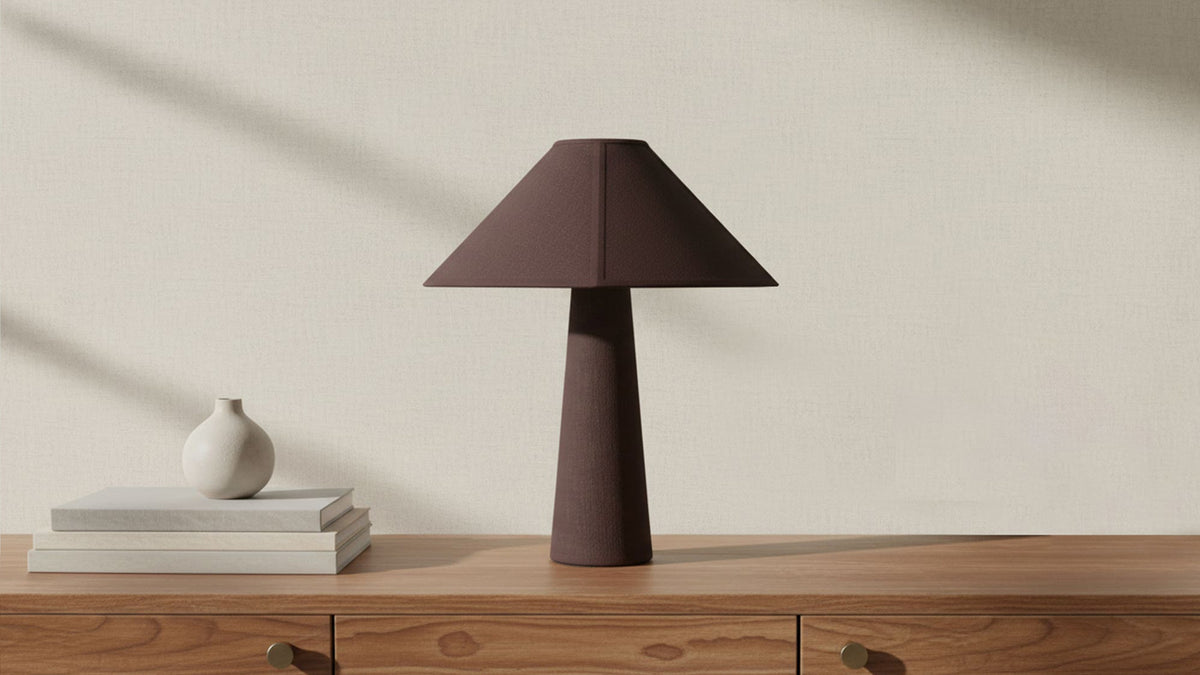 The Gigi table lamp in dark brown linen and cotton on a walnut console, its tapered base and geometric shade reading as a dark silhouette against a pale plaster wall