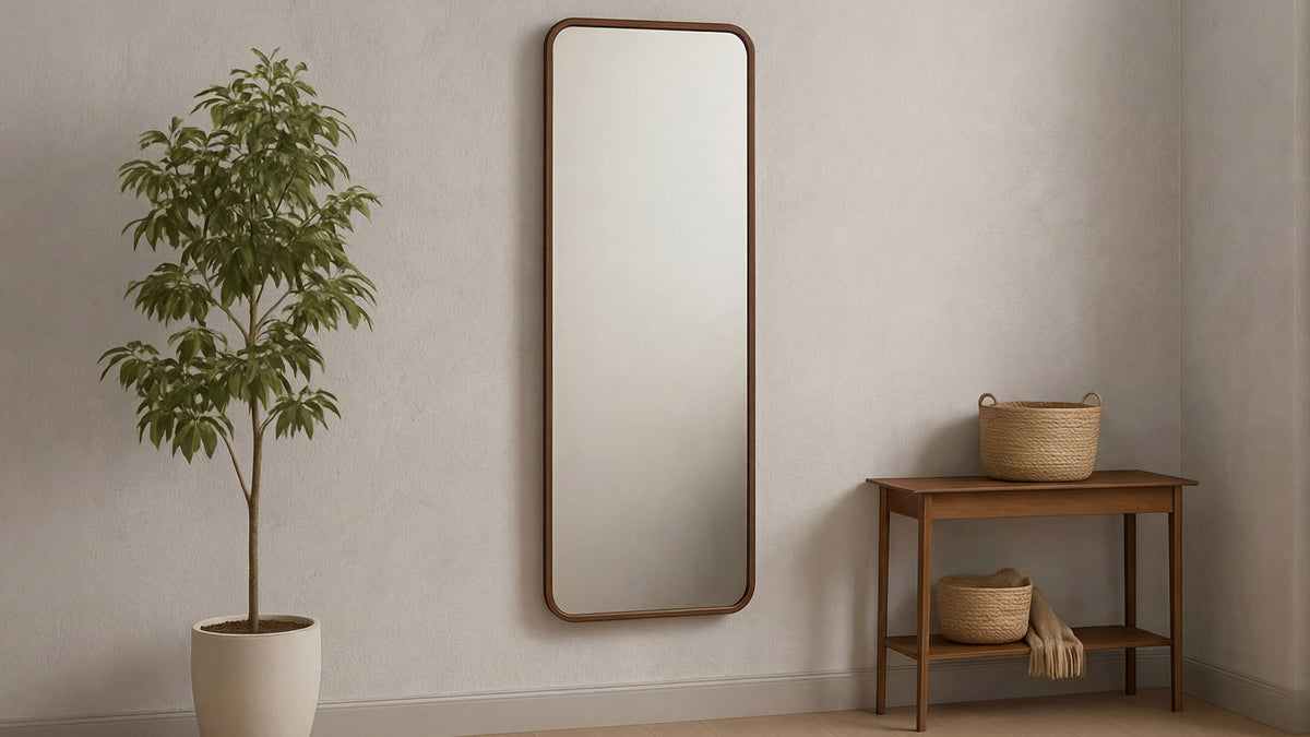 The Kai floor mirror in walnut on a pale wall between a potted tree and a small wooden bench holding woven baskets