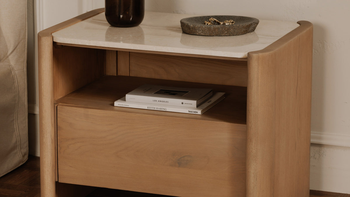 Three-quarter view of the Moe's Home Collection Lane nightstand in natural oak, two books stacked in the open cubby below the marble top and a stone dish of jewellery above