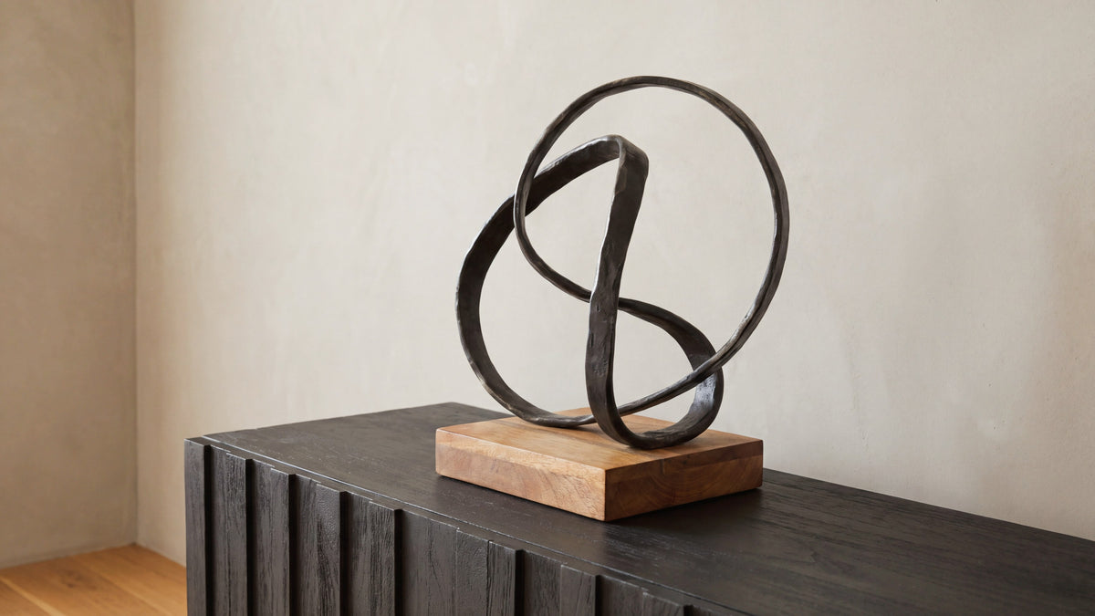 The Lida sculpture on a black fluted console, its looping aged-black aluminium ribbon rising from a mango wood base against a pale wall