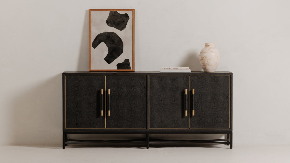 Moe's Home Collection Mako sideboard in black shagreen-patterned polyurethane on a black metal base, its brass-capped pulls and brass door trim against a pale wall, with a framed abstract print leaning behind