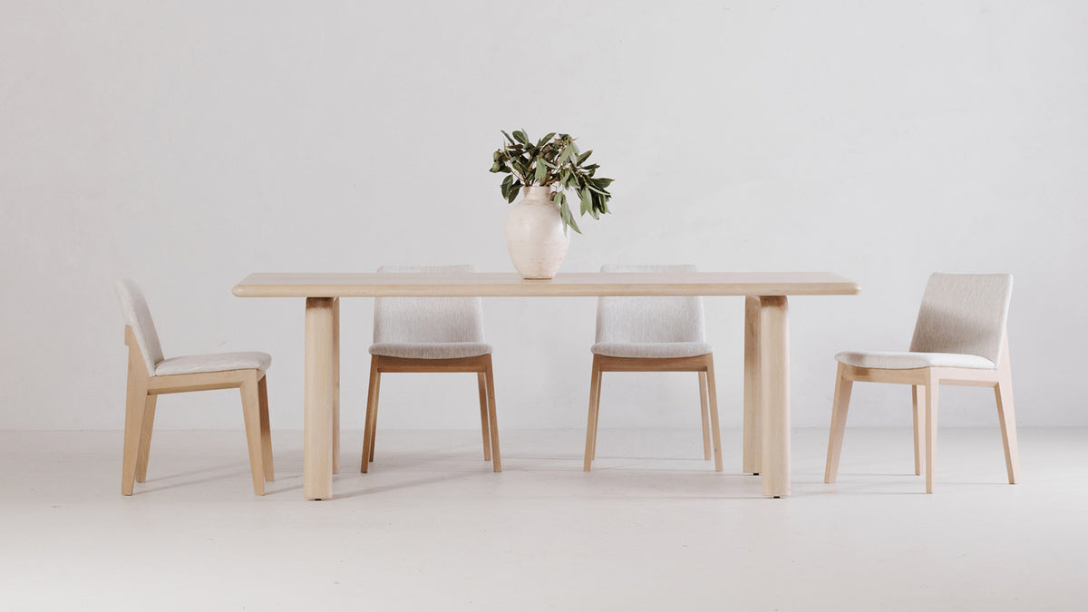 Moe's Home Collection Malibu dining table in solid white oak, set with four pale upholstered dining chairs and a cream ceramic vase of olive branches against a plain studio wall