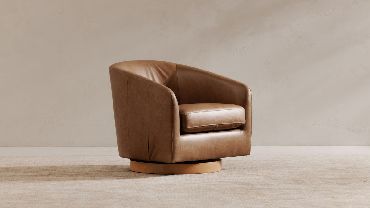 A Moe's Home Collection Oscy swivel chair in tan top-grain leather on a pale ash swivel base, shown at three-quarter angle on a woven rug against a warm plaster wall