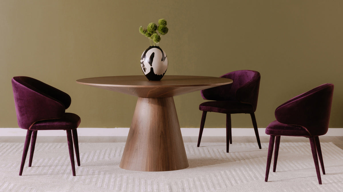 Moe's Home Collection Otago round dining table in walnut against an olive green wall, standing on a cream textured rug with three plum velvet chairs drawn up to it and a black and white ceramic vase of green blooms on the top