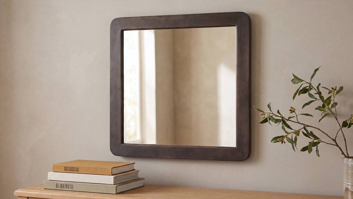 The square Perth mirror on a sunlit wall at a slight angle, a stack of books below it and an olive branch crossing the frame