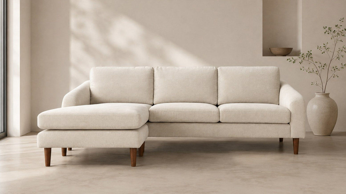 Moe's Home Collection Quinn Sectional in oatmeal with the chaise ottoman on the left, photographed straight on in a pale plaster room with a full-height window at the left, a recessed niche holding a wooden bowl, and a branch in a cream vase at the right