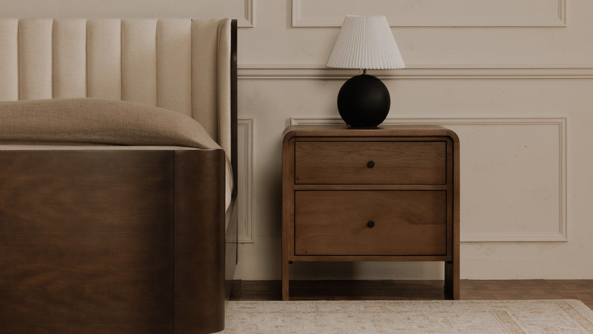 Moe's Home Collection Riley nightstand in oak beside a channel-tufted bed in a panelled room, its two drawers and small black knobs below a waterfall edge, with a round black lamp and pleated shade on top