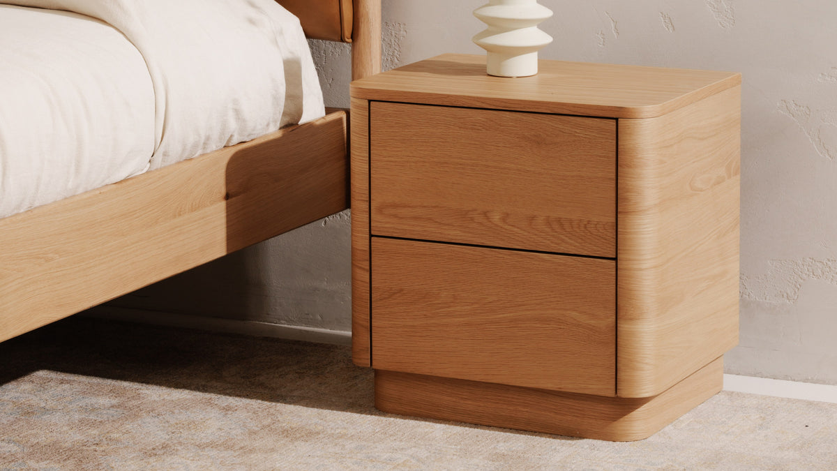 Moe's Home Collection Round Off Tall nightstand in light oak beside an oak bed, its two handleless drawers separated by fine shadow lines, the rounded case standing on a recessed plinth with a white sculptural vase above