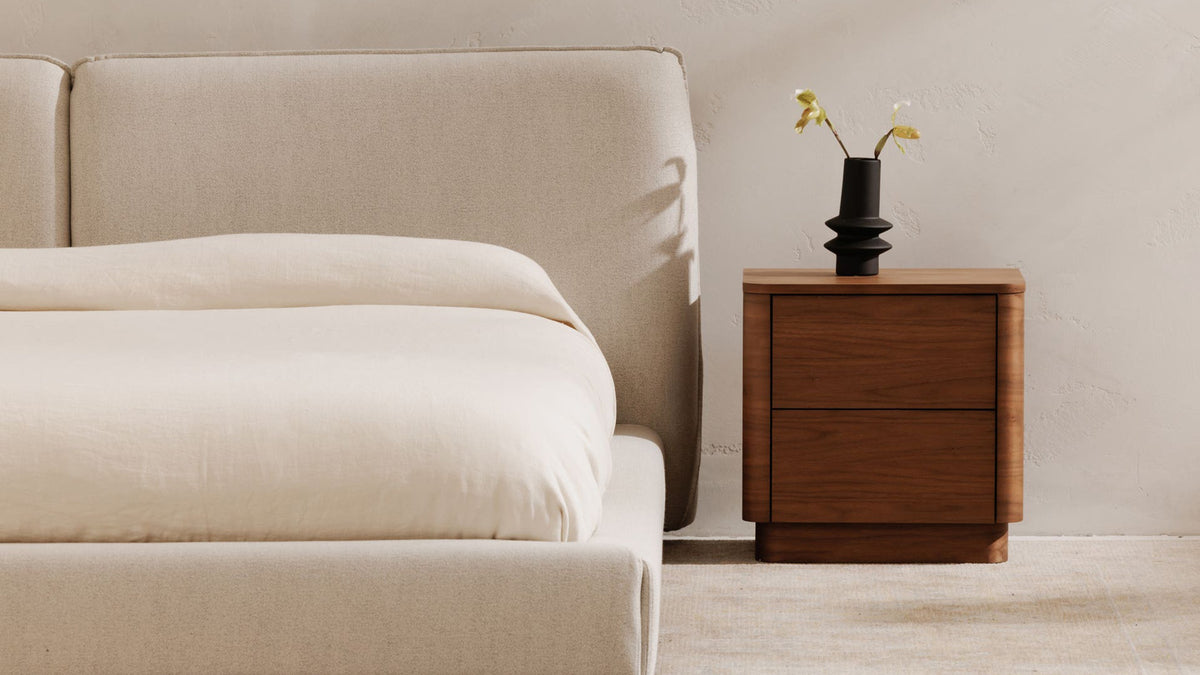 Moe's Home Collection Round Off Tall nightstand in walnut brown against a plaster wall beside a cream upholstered bed, two handleless drawers in a softly rounded case on a recessed plinth, with a black ribbed vase on top