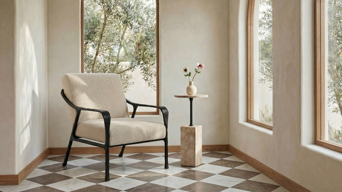 The Moe's Home Collection Rowe accent chair in beige textured polyester on a black hammered cast-aluminum frame, on a checkered marble floor beside a stone pedestal side table holding a small vase of anemones, in a plaster room with arched windows onto an olive grove
