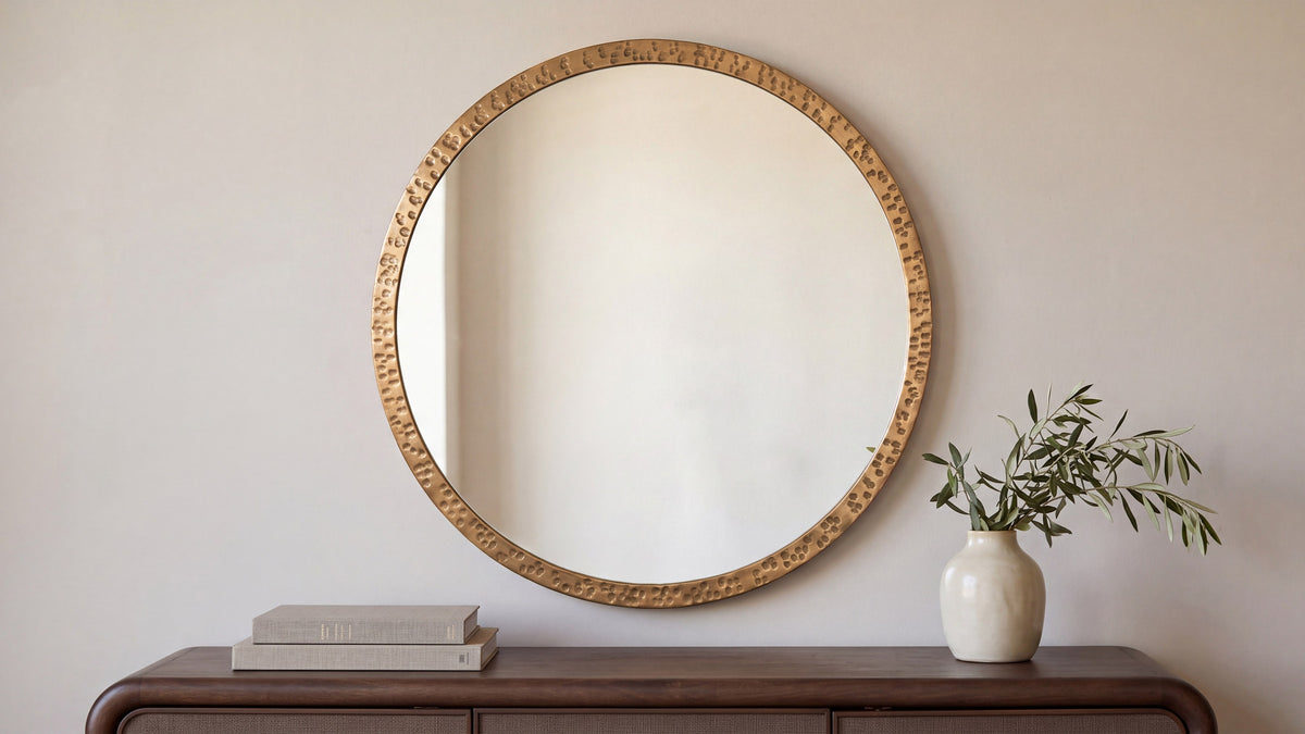 The large Royce mirror in bronze above a dark wood console, the hammered rim catching warm light, with books and a pale vase below