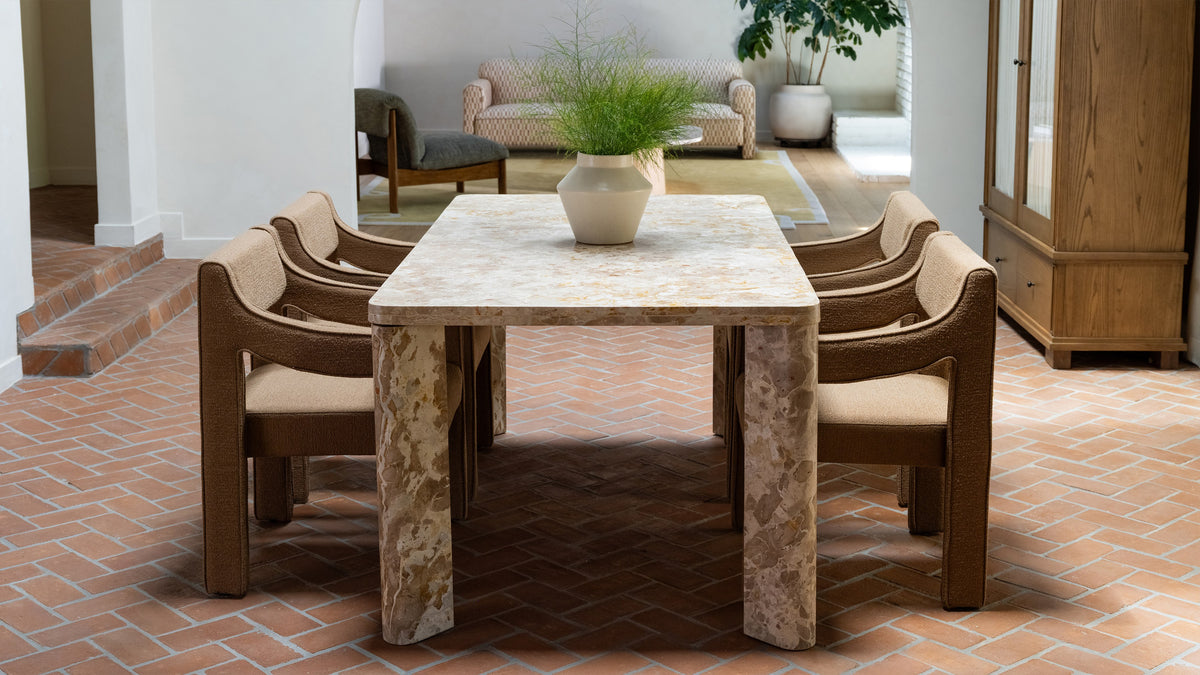 Moe's Home Collection Segment dining table in beige and gold Panther marble, its four angled legs standing on a terracotta brick floor, surrounded by brown boucle armchairs in a white plaster room
