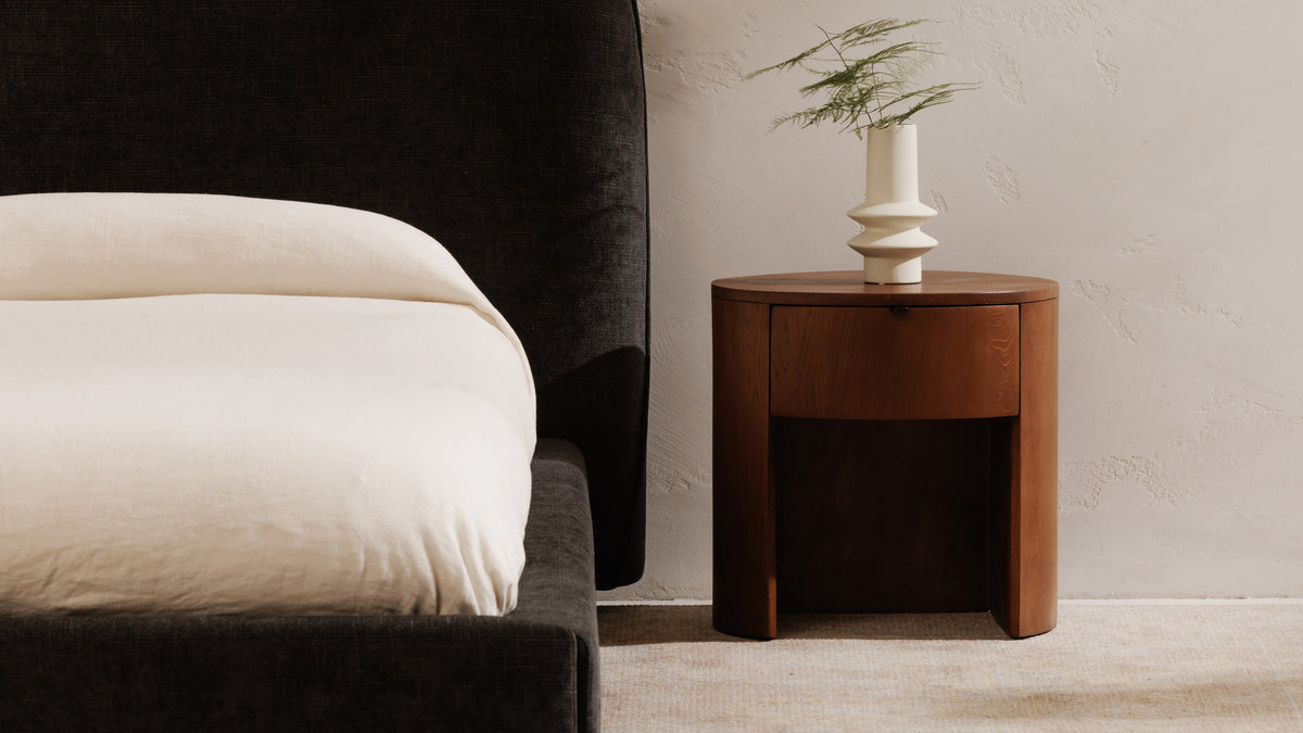 Moe's Home Collection Theo 1-Drawer nightstand in brown oak beside a charcoal upholstered bed, its single curved drawer sitting above an open recess between the drum's two sides, with a white sculptural vase on top