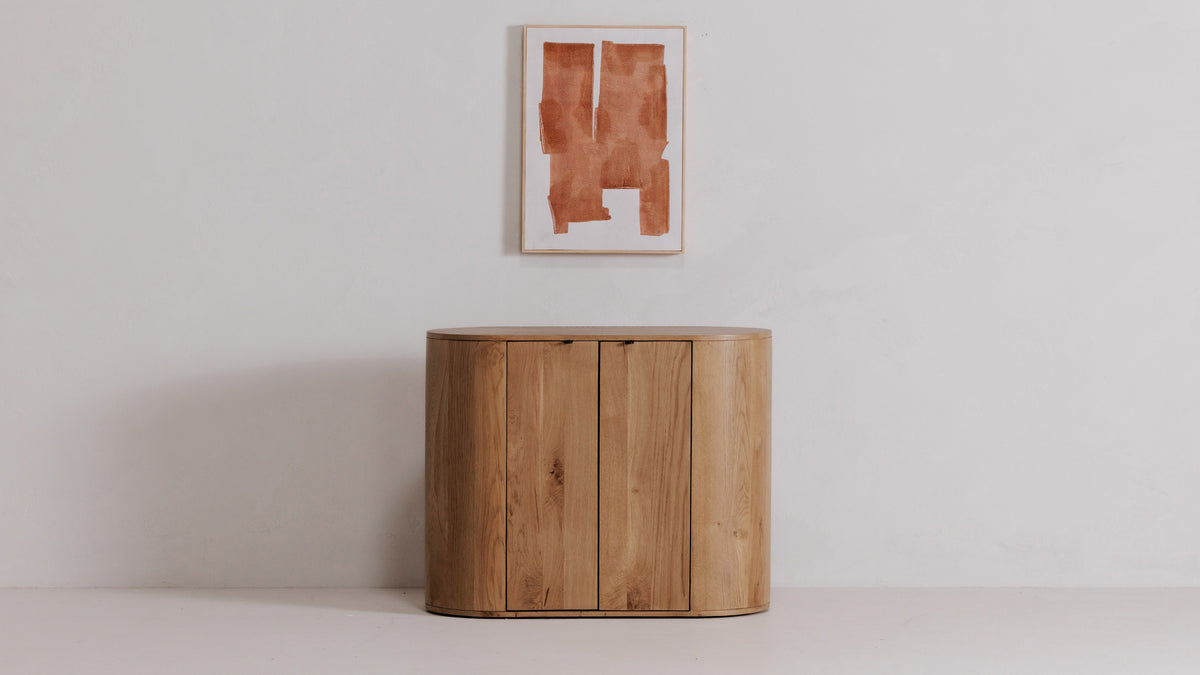 Moe's Home Collection Theo oak cabinet in natural oak, an oval two-door cabinet standing alone beneath a framed print
