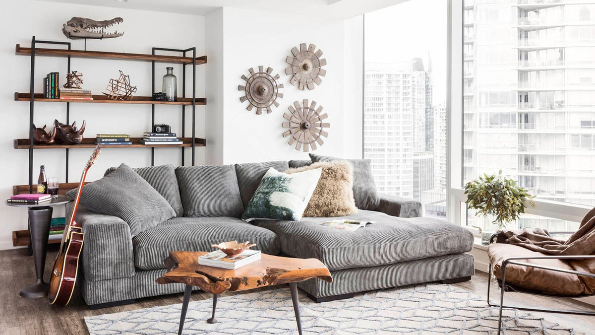 Moe's Home Collection Vancouver Large bookshelf, a black iron frame carrying five solid acacia shelves of books and objects, standing behind a grey sectional in a city apartment