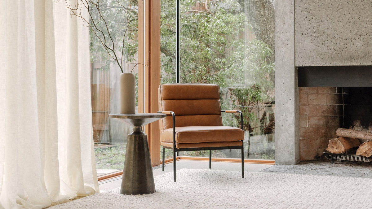 A Moe's Home Collection Verlaine armchair in chestnut brown top-grain leather on a slim black metal frame, on a cream wool rug beside a floor-to-ceiling window, with a bronze pedestal side table and a board-formed concrete fireplace to the right