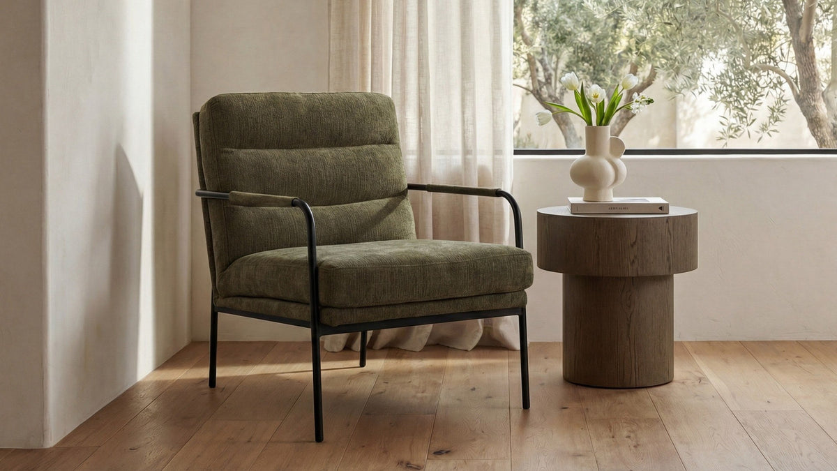 A Moe's Home Collection Verlaine armchair in olive polyester on a powder-coated iron frame, on a wide-plank oak floor against a plaster wall, with an oak drum side table holding white tulips and an olive tree through the window behind