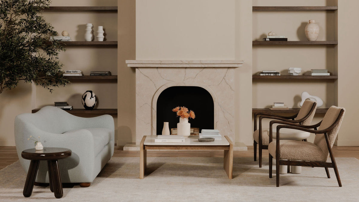 A pair of Moe's Home Collection Yates accent chairs in beige faux shearling on deep brown solid oak frames, angled toward each other beside a limestone fireplace, with a pale curved sofa and a marble-topped oak coffee table opposite on a cream rug