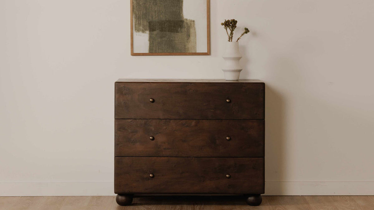 Moe's Home Collection York 3 Drawer nightstand in dark brown burl against a pale wall, its three drawers each carrying two small brass knobs, standing on rounded acacia ball feet below a framed green abstract painting with a white vase of dried seedheads on top