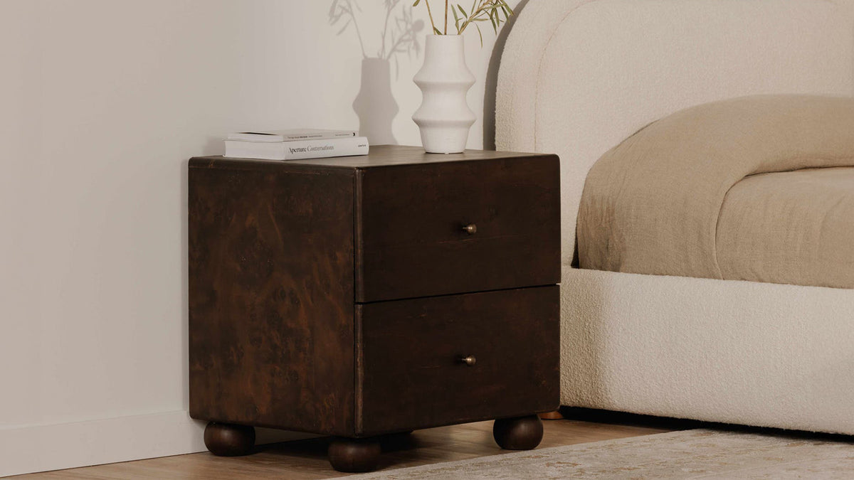 Moe's Home Collection York nightstand in dark brown burl beside a cream boucle bed, its two drawers and small brass knobs above four rounded acacia ball feet, with a white ribbed vase and a stack of books on top