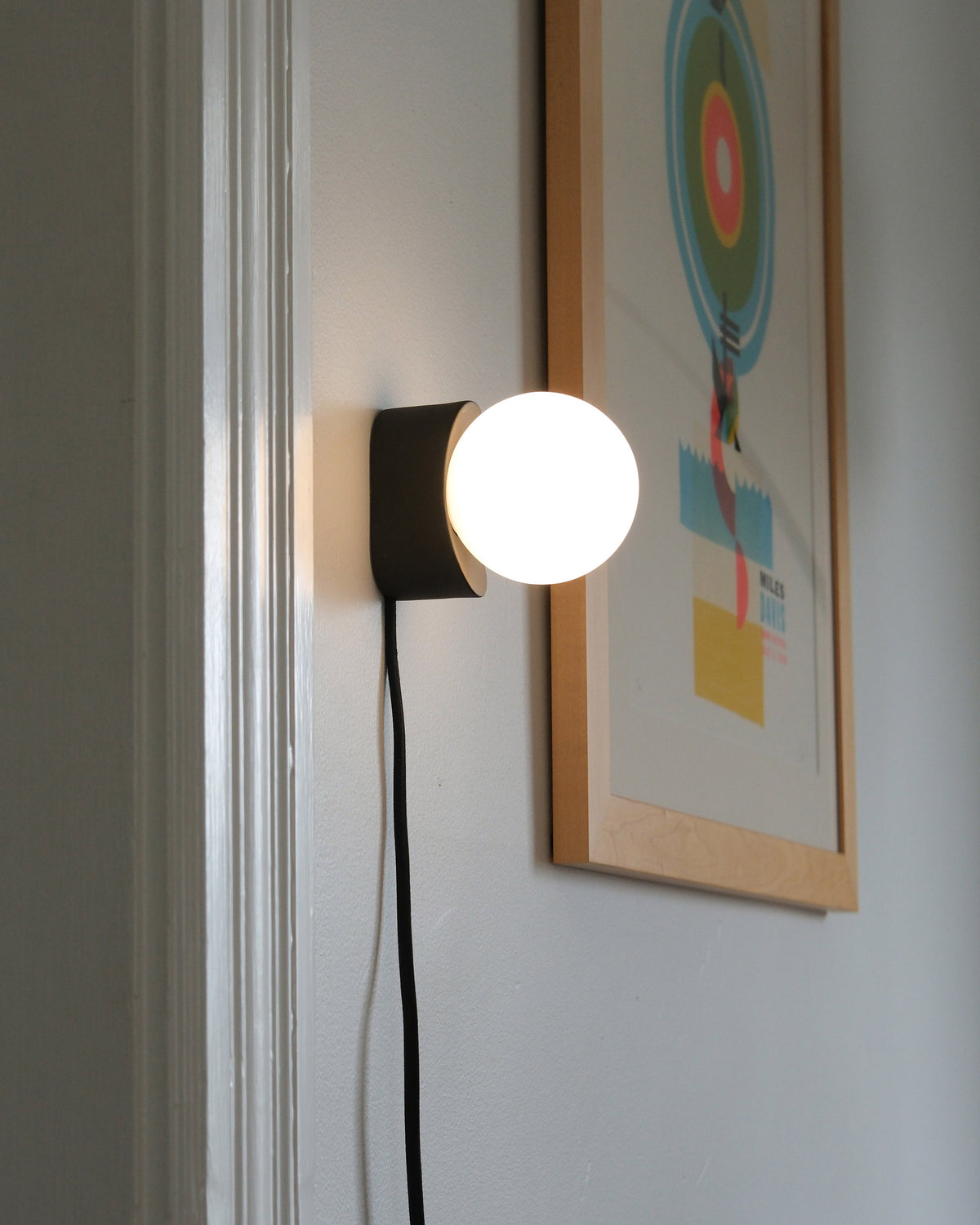 Iota Sconce in Black, lit, beside a framed print