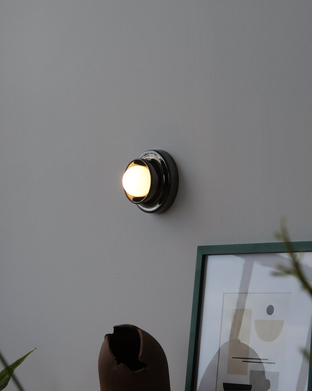 Q Sconce in Chrome, lit, beside framed art