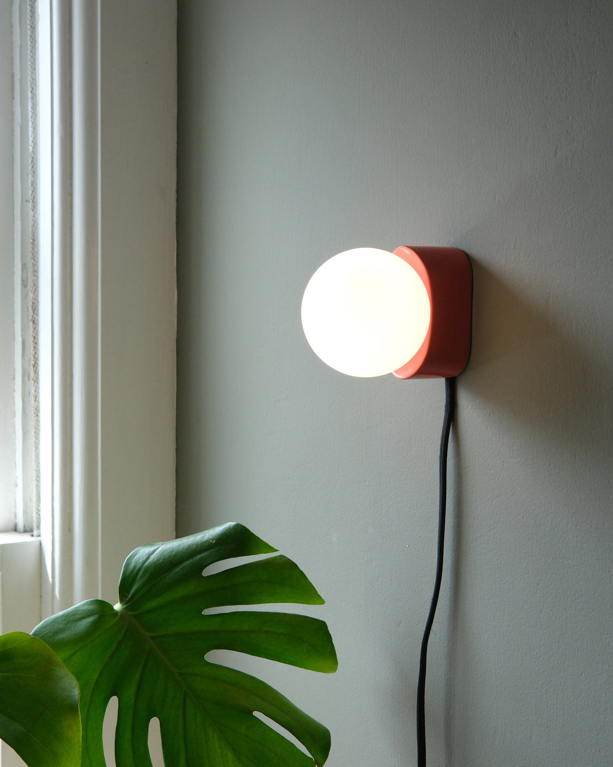 Iota Sconce in Vermilion, lit, beside a window