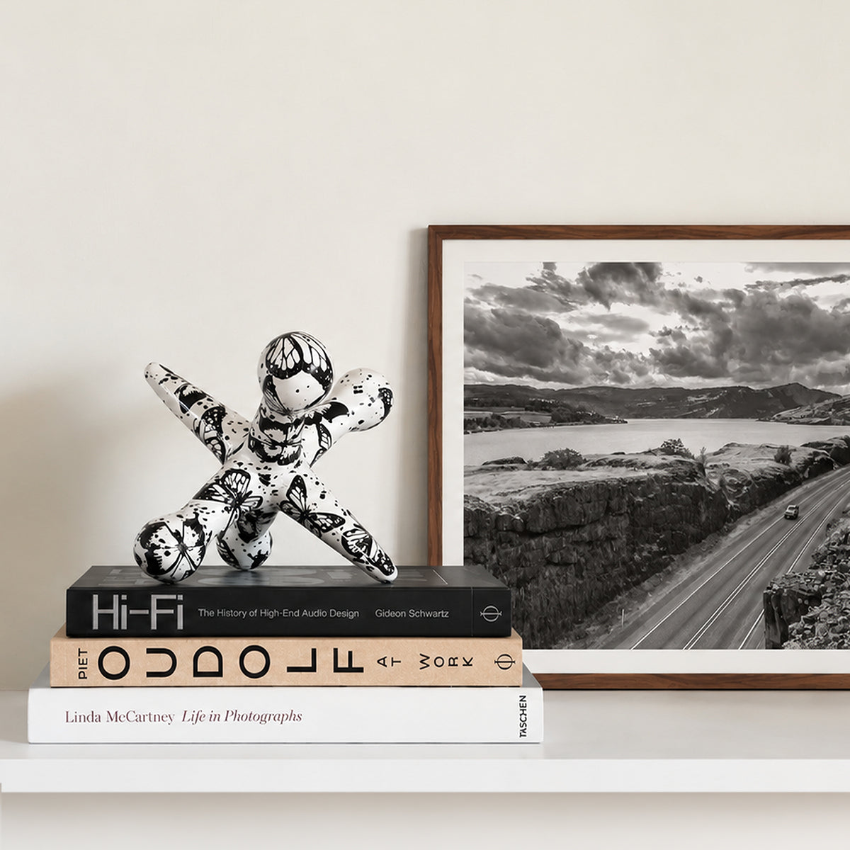 A framed black and white landscape photograph leaning against a wall on a white shelf, next to a small black-and-white patterned figurine resting on stacked books.