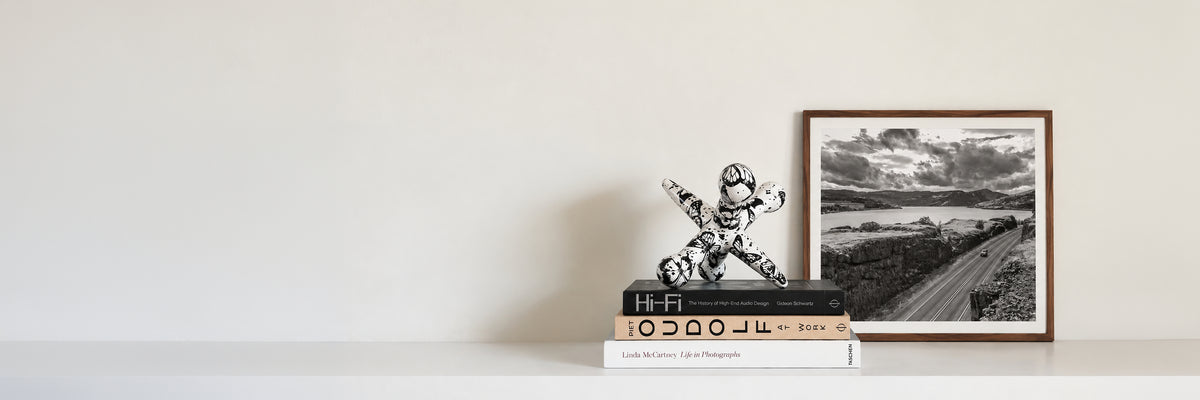 A framed black and white landscape photograph leaning against a white wall on a white shelf, next to a small white figurine and stacked books.