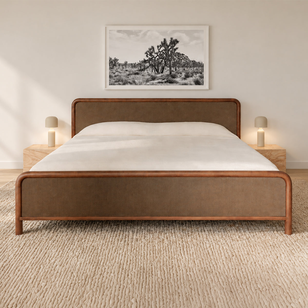 A modern platform bed with a warm brown upholstered headboard and footboard, styled in a minimalist bedroom with a white mattress, wooden nightstands, and a framed black-and-white landscape print on the wall.