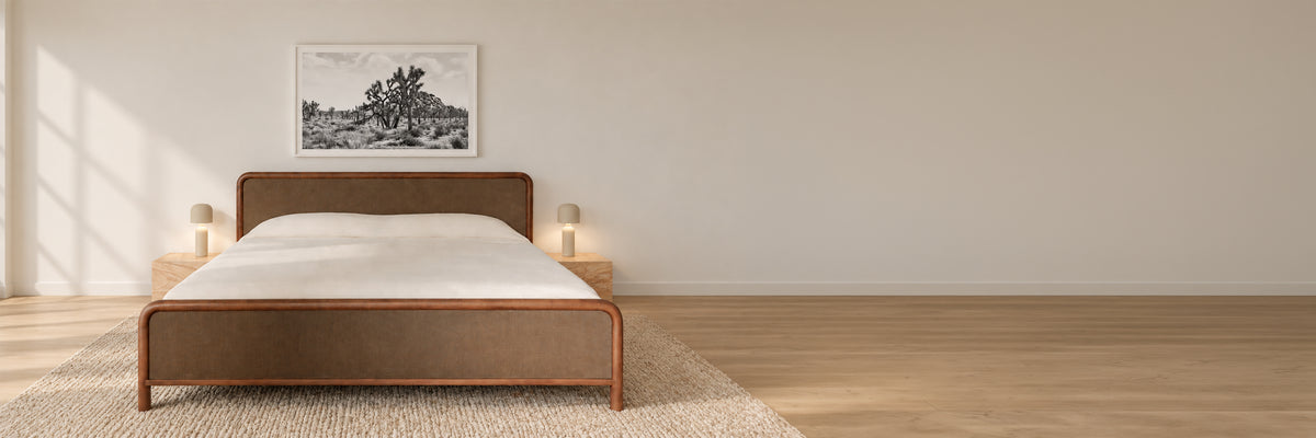 A minimalist bedroom scene featuring a low-profile bed with a brown upholstered headboard and footboard, dressed in white bedding. The bed is positioned on a light beige rug against a warm beige wall, with a black and white landscape print hanging above it.