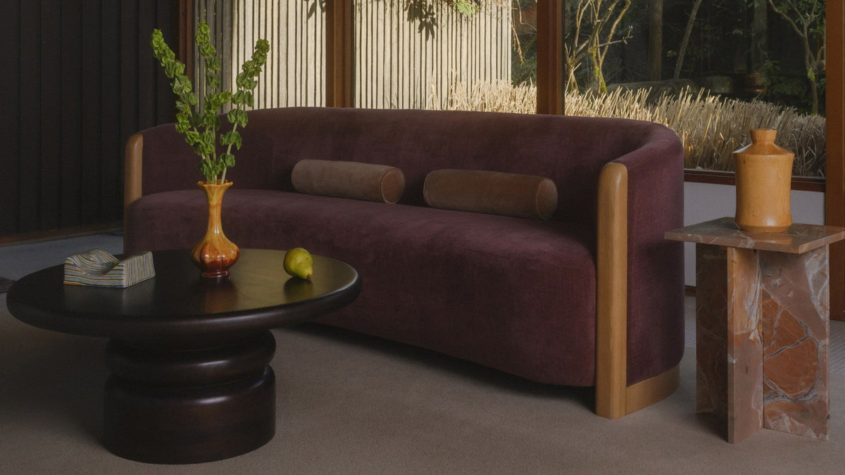 Moe's Home Collection Anya sofa in berry chenille with two bolster cushions, its oak corner post and base rail catching low evening light in a dark timber room beside a round black table and a stone side table