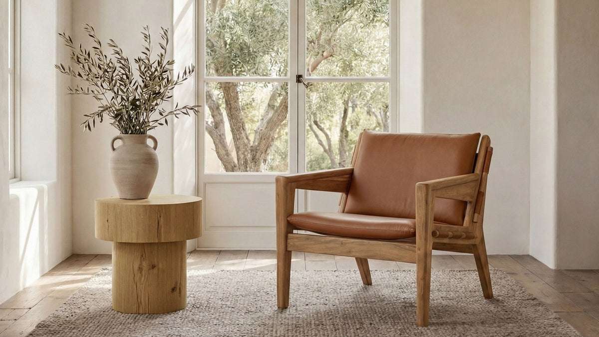 The Moe's Home Collection Baker chair in brown top-grain buffalo leather on a pale acacia frame, set on a woven rug beside a round oak side table holding a stoneware vase of olive branches, in front of white French doors opening onto an olive grove