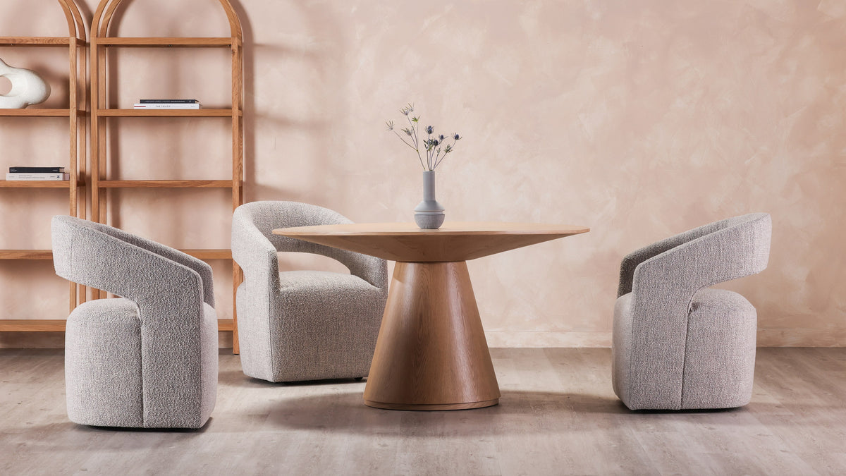Three Moe's Home Collection Barrow rolling dining chairs in gray storm boucle around a round oak pedestal table, against a pink plaster wall with an arched oak shelving unit