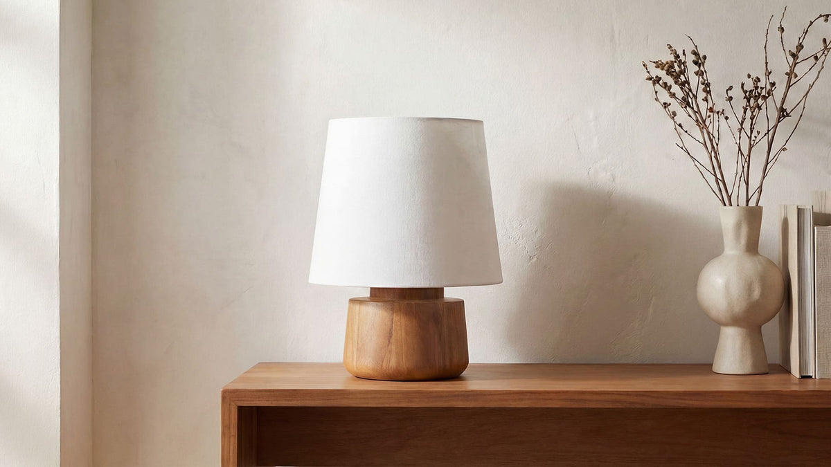 The Bibi table lamp on a walnut console against a plaster wall, its rounded solid mango base under a pale fabric shade, beside a sculptural ceramic vase holding dried stems