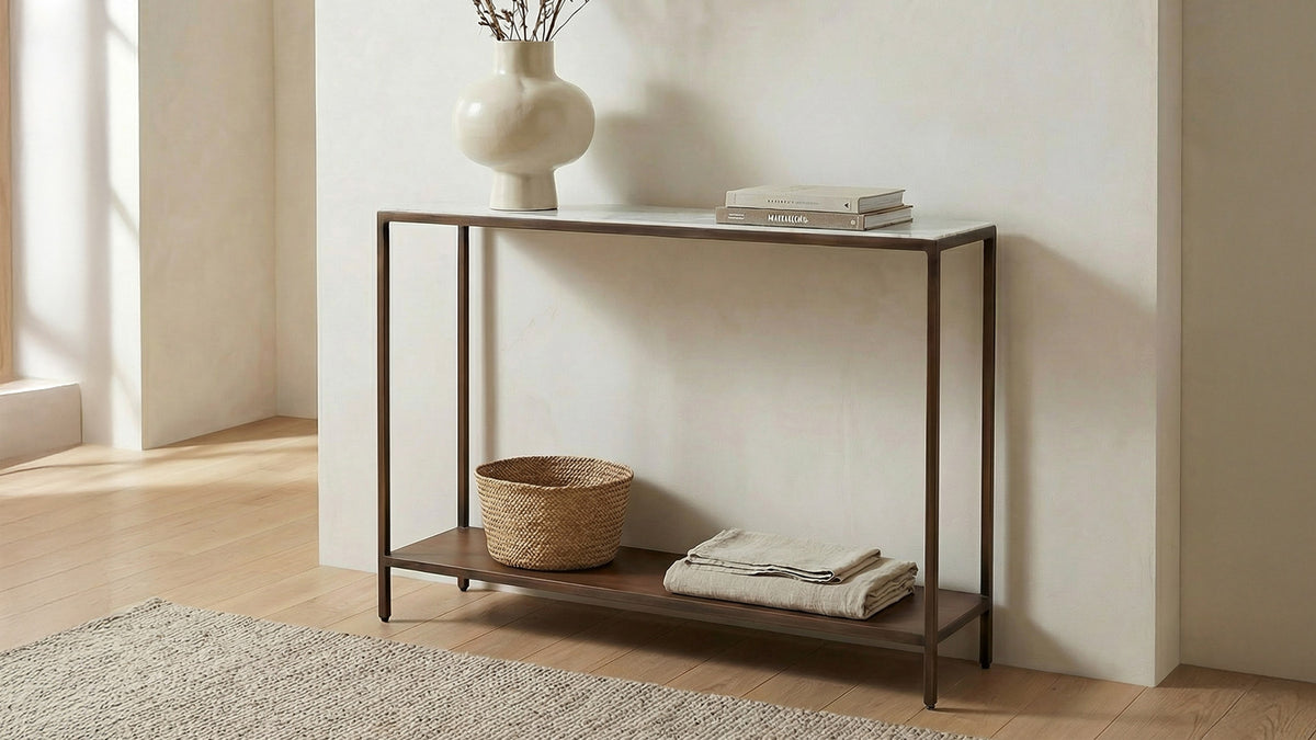 Moe's Home Collection Bottego console table in antique copper with a white marble top, styled with a ribbed vase of dried stems and a stack of books, a woven basket and folded linens on the lower shelf