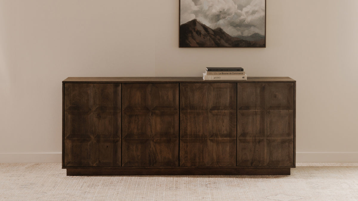 Moe's Home Collection Bowen sideboard in deep brown mango wood against a pale wall, its blocky door fronts catching low light, with a framed landscape above and a stack of books on top