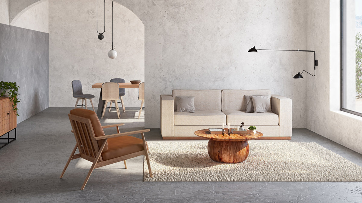 Moe's Home Collection Bradbury round coffee table in Natural acacia, its spherical pedestal base on a cream rug in a sunlit plaster-walled living room with a pale sofa and a leather lounge chair