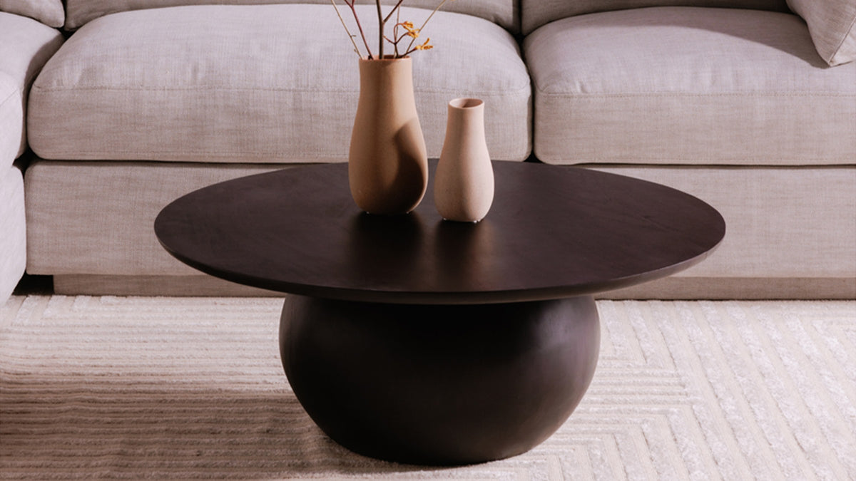 Moe's Home Collection Bradbury round coffee table in Black acacia, its dark spherical base and low round top seen close against an oatmeal sectional, holding two clay vases
